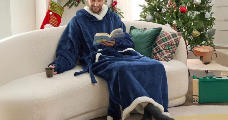 man wearing a blue wearable blanket hoodie reading a book on a sofa, Christmas decor and Christmas tree in the room behind him