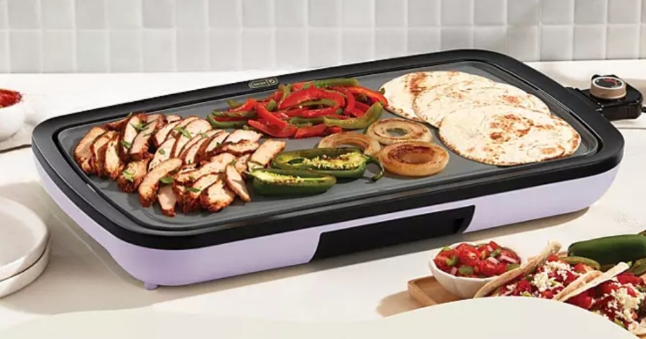 Dash griddle with a black ceramic cooktop and lavender purple base with food for fajitas cooking on it