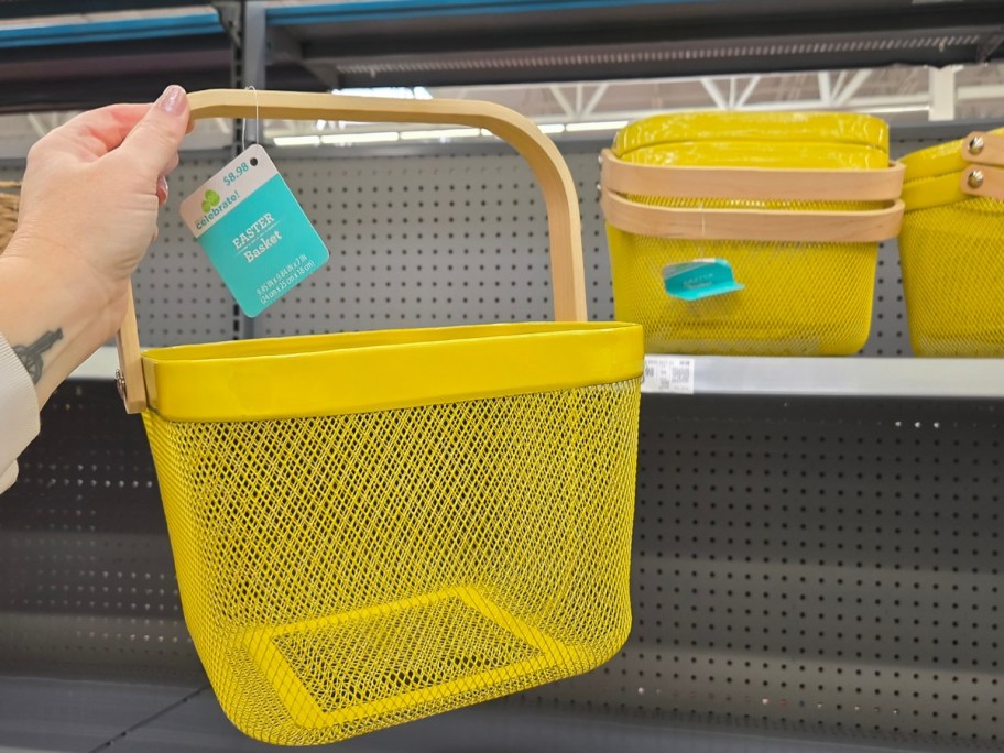 hand holding a yellow mesh wire basket with a wood handle, more baskets on a store shelf behind it