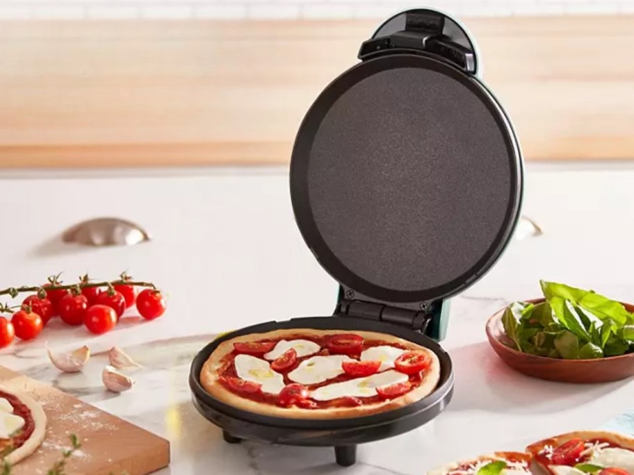 Dash round griddle open with cooked mini pizza inside next to pizza ingredients on a kitchen counter