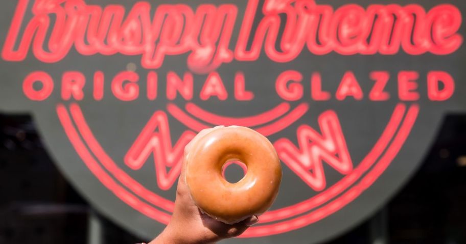 Hand holding up s Krispy Kreme Glazed doughnut in front of the hot & ready now sign