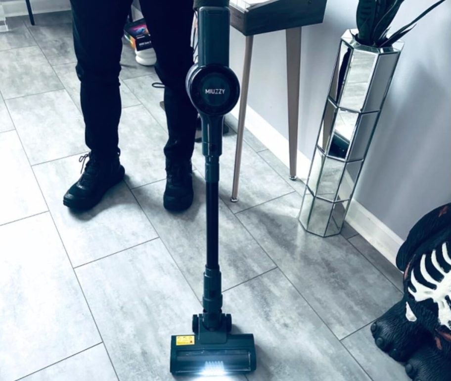 A MIUZZY Cordless Vacuum Cleaner, (1) next to a man's feet using the vacuum