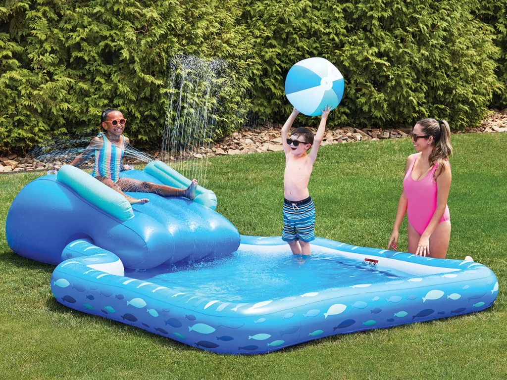 two kids playing in blue inflatable pool with slide and mom kneeling next to them