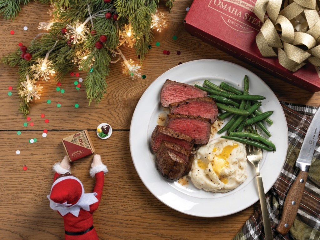 Plate of steak, mashed potatoes and green beans next to little elf with miniature steak box