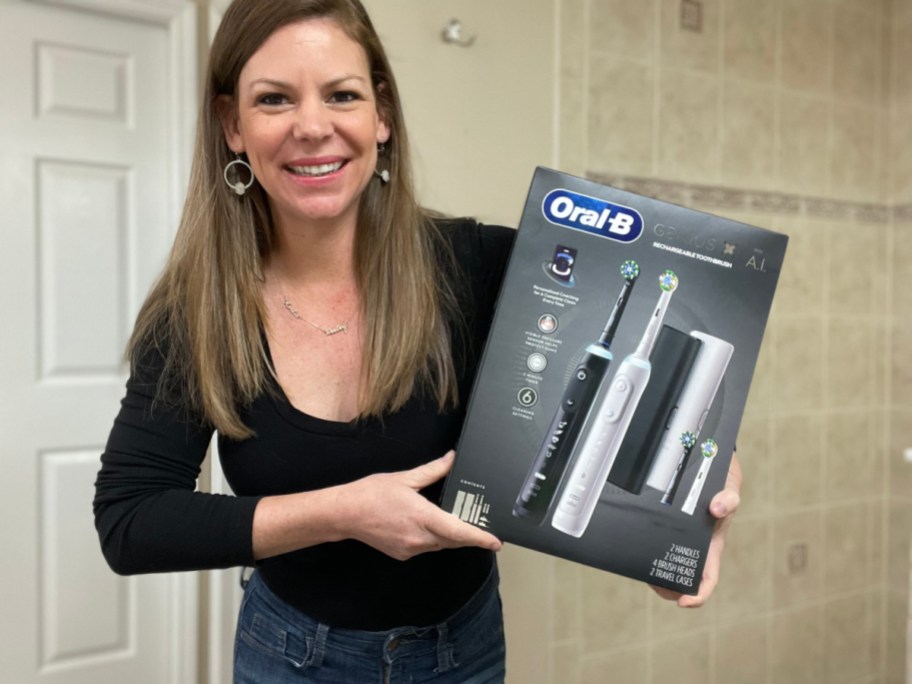 smiling woman holding an oral b toothbrush package standing in a bathroom