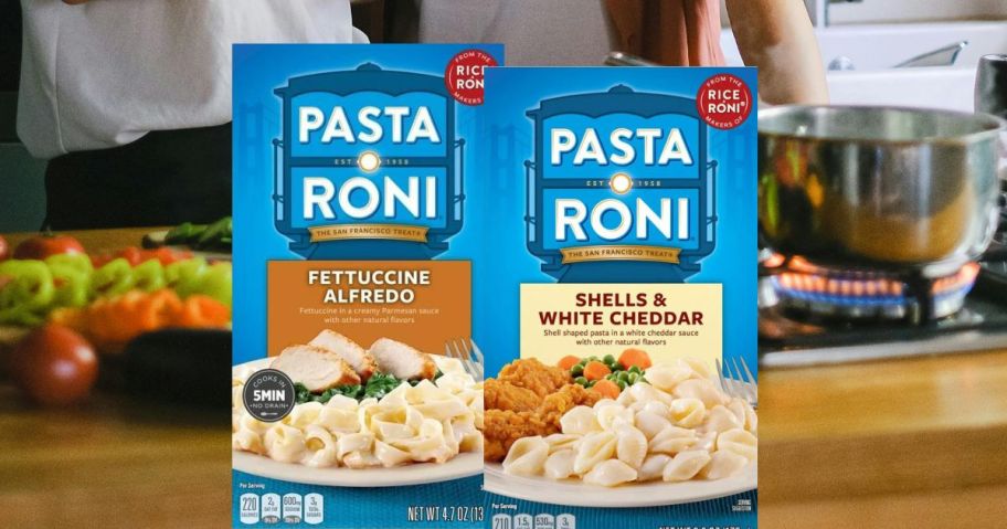 Two boxes of Pasta roni in front of a counter of veggies and a pot of boiling water