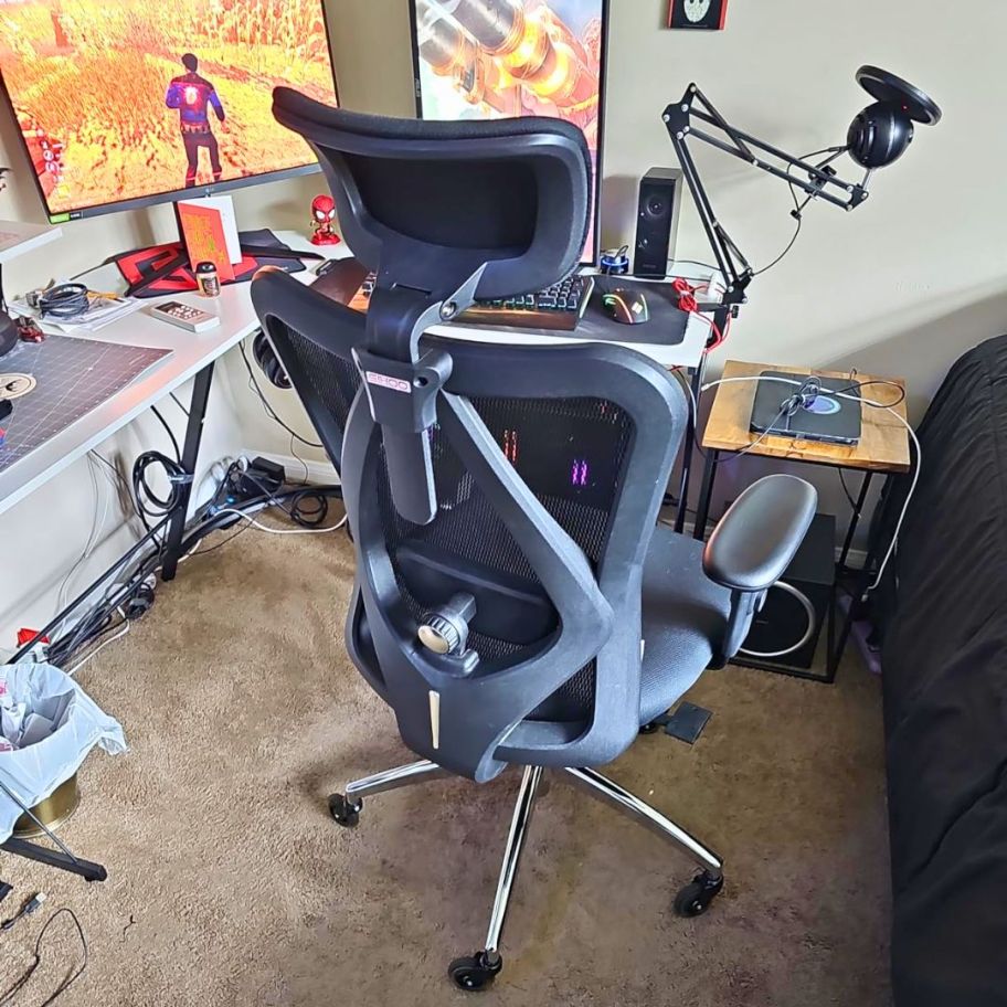 back of a black mesh office chair with headrest and armrests sitting next to a desk with monitors and gaming equipment