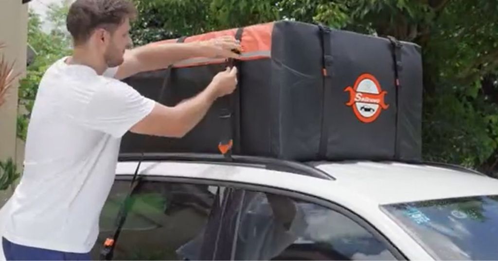 man installing a black and orange car rooftop carrier bag on a white SUV