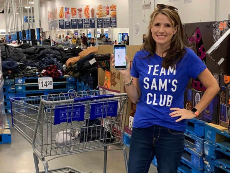 woman wearing a Team Sam's Club T-Shirt while holding up her phone next to a Sam's Club shopping cart