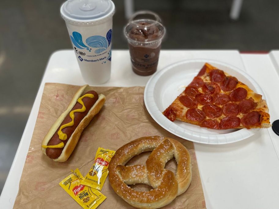 A table filled with Sam's Club food items including a fountain soda, ice cream, a hot dog, a slice of pepperoni pizza and a soft pretzel