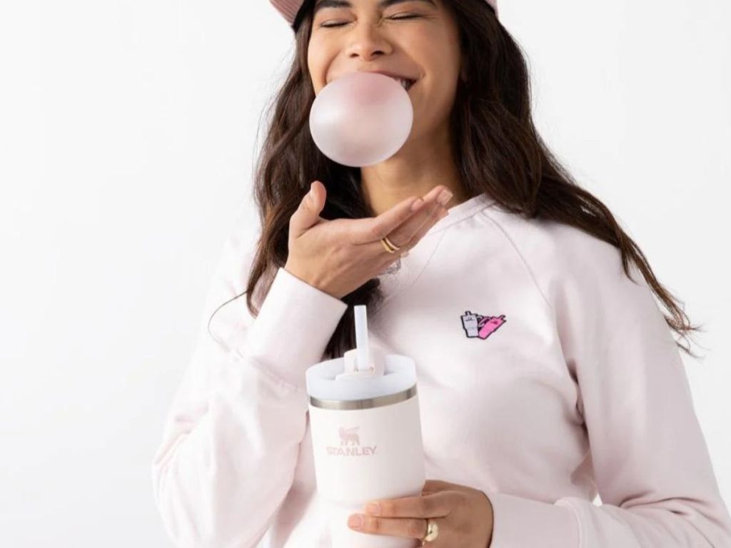 A woman blowing a bubble, holding a Stanley cup, and wearing a Stanley sweatshirt 