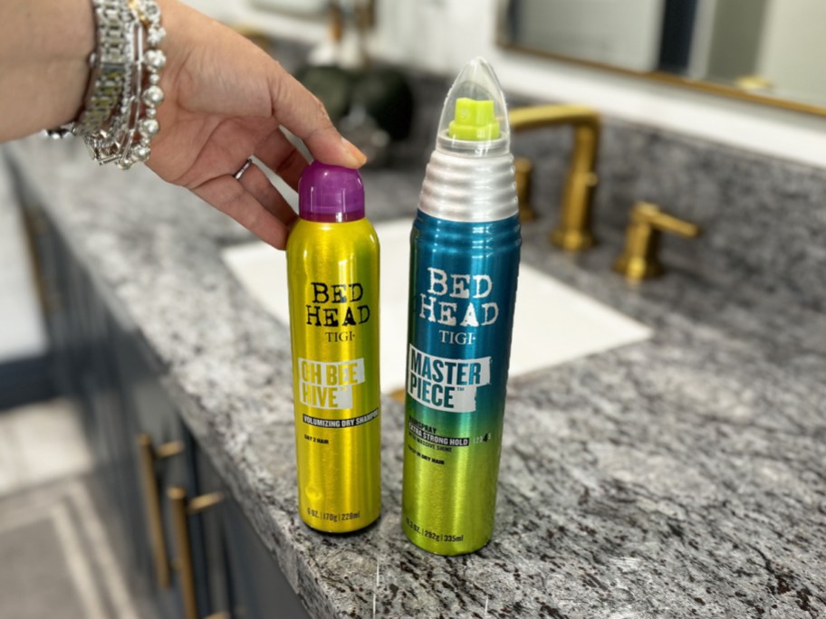 two TIGI Bed Head Sprays on bathroom counter with hand touching one