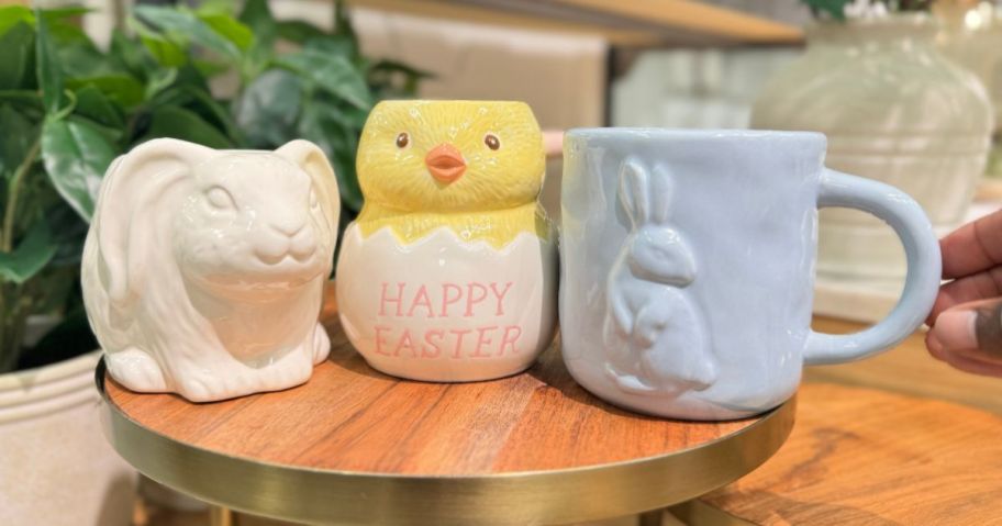 Three ceramic easter mugs displayed on a round wooden riser - featuring white rabbit,, happy easter chick and blue bunny 