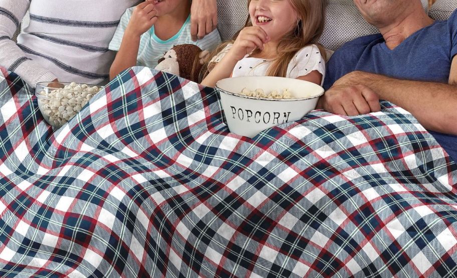 a family of four under a family size blanket on a sofa sharing a bowl of pop corn