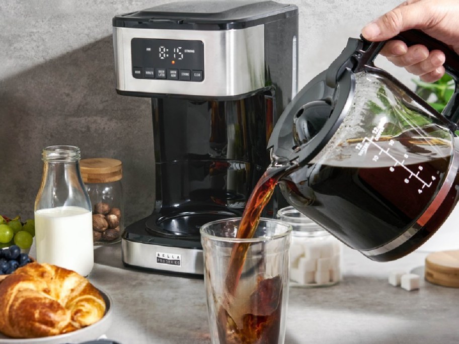 coffee maker in the background while hand is pouring coffee from a carafe into a glass cup