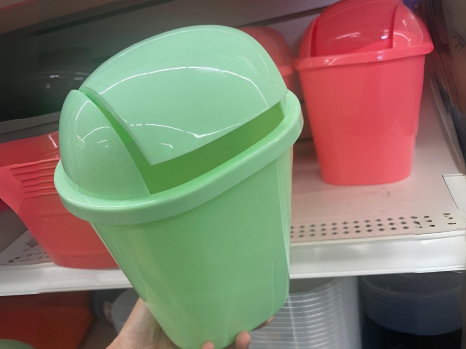 hand holding a small plastic green trash bin with a flip lid, more behind it in reddish orange