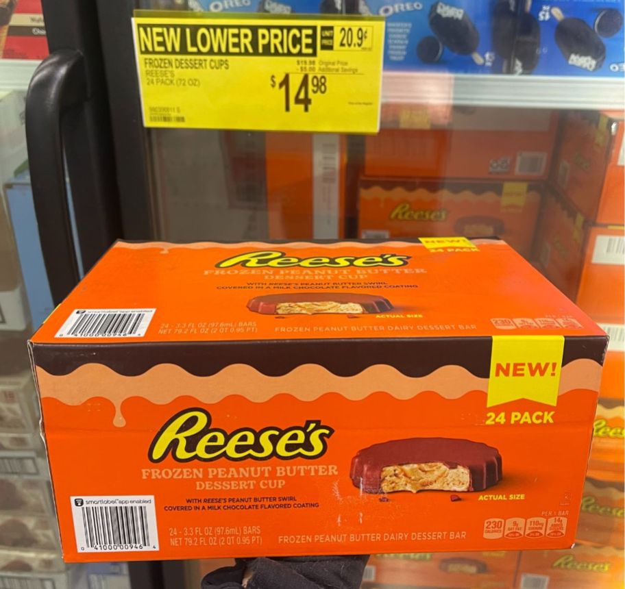a box of reeses frozen peanut butter cups held up in front of a store freezer door below a pricing sign showing the price drop