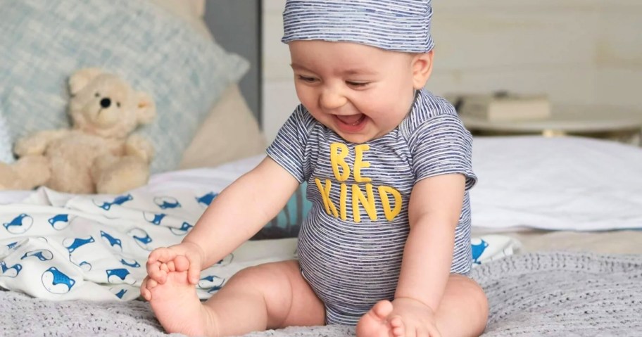 baby boy sitting on a bed wearing a blue striped onesie that says "Be Kind" in yellow and a matching hat