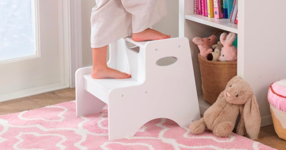 child standing on white stool near bookshelf on pink rug
