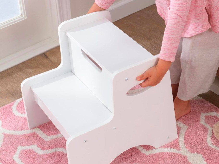 child lifiting white wooden stool off pink rug on floor 