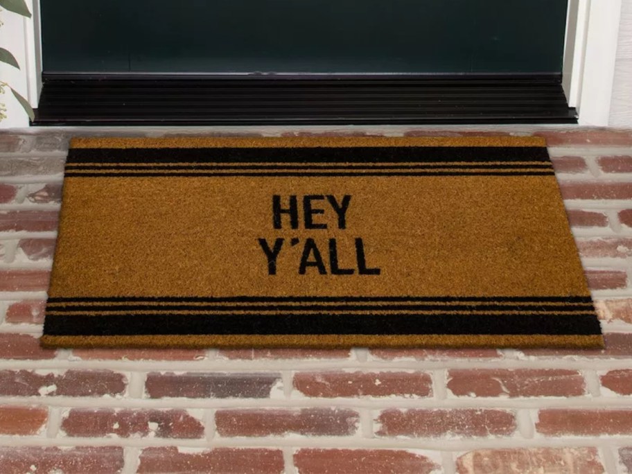 tan and black coir doormat that says "Hey Ya'll" on brick in front of a door