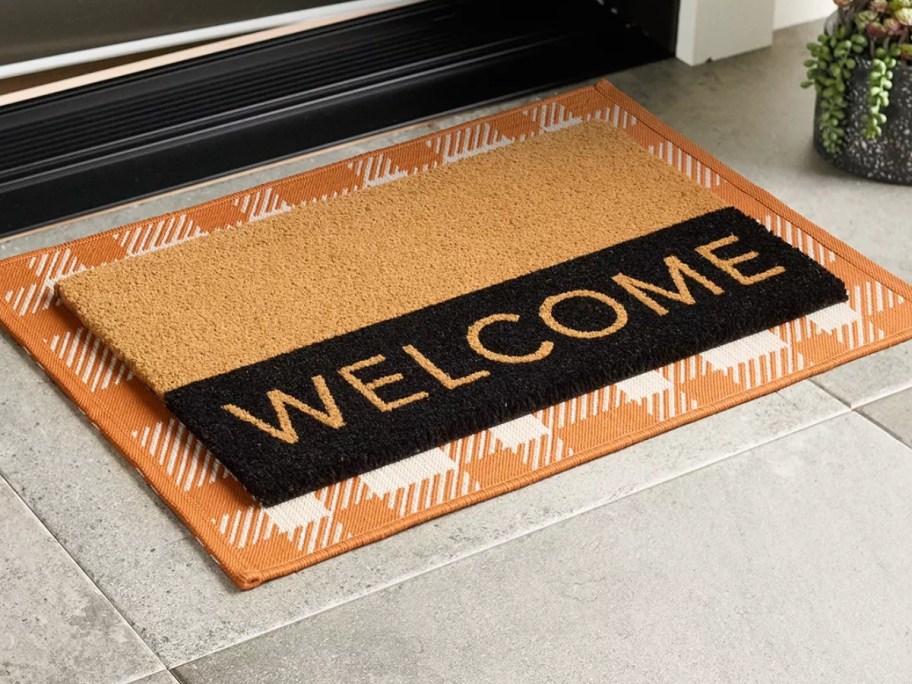 tan and black coir doormat with "Welcome" laying on an orange plaid rug in front of a door
