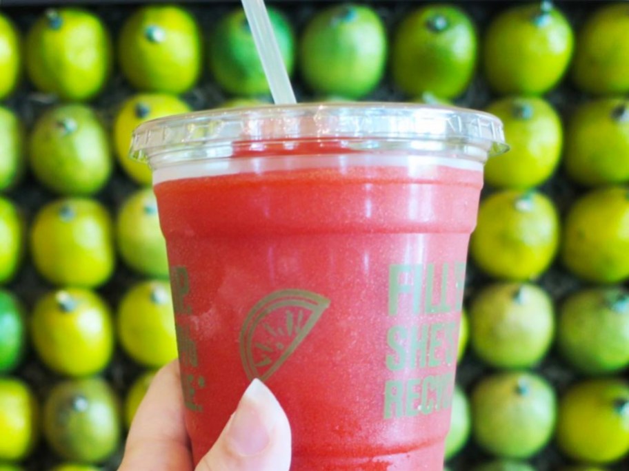 holding a frozen red drink in front of a wall of limes
