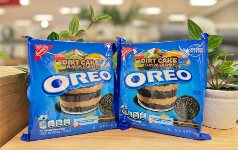 two packages of new dirt cake flavor oreos side by side on a wooden surface in front of foliage