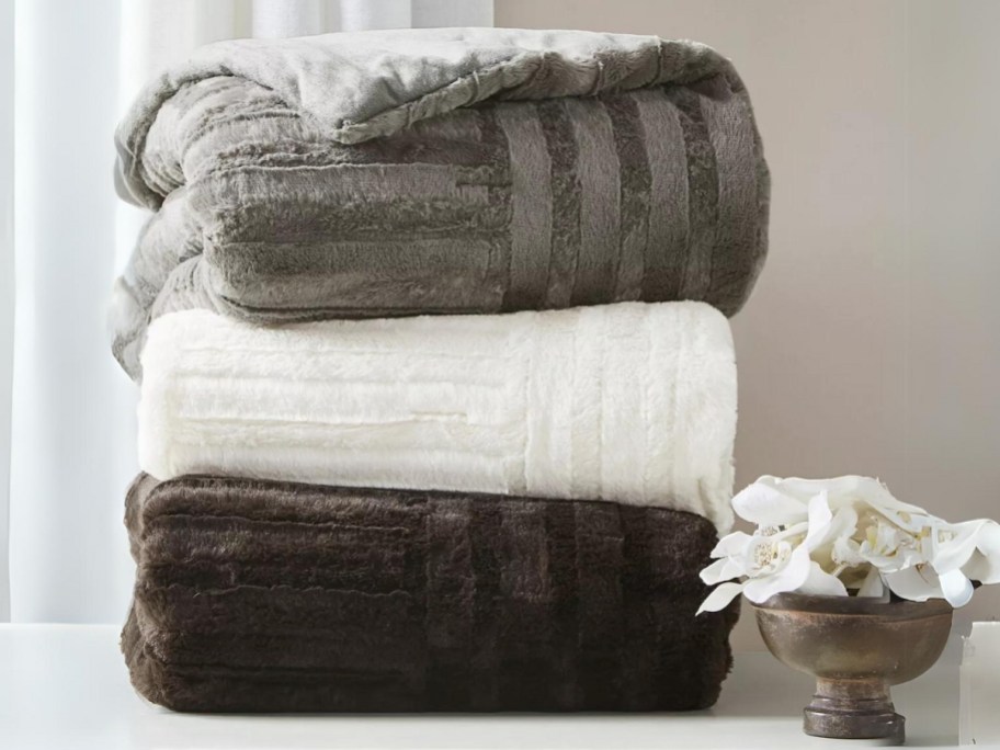 three thick faux fur blankets in grey, white and brown stacked on a bench