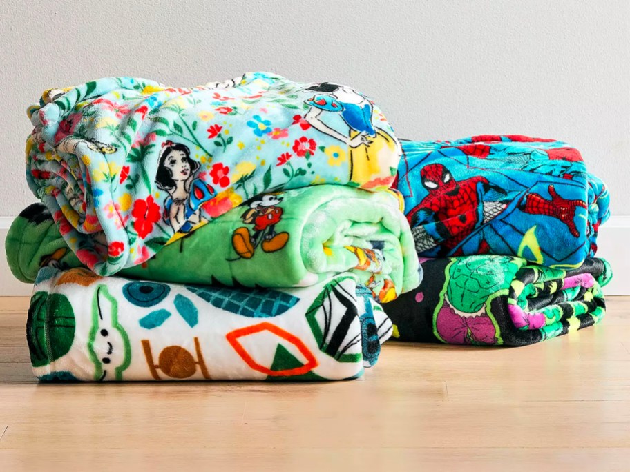 the big one disney blankets folded and stacked on floor featuring snow white, mickey, grogu, spiderman and the hulk