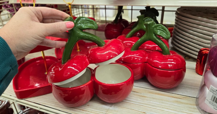 A hand lifts a ceramic cherry-shaped container with a green stem lid, among other similar bright red containers on a wooden shelf. Stacks of plates are nearby.