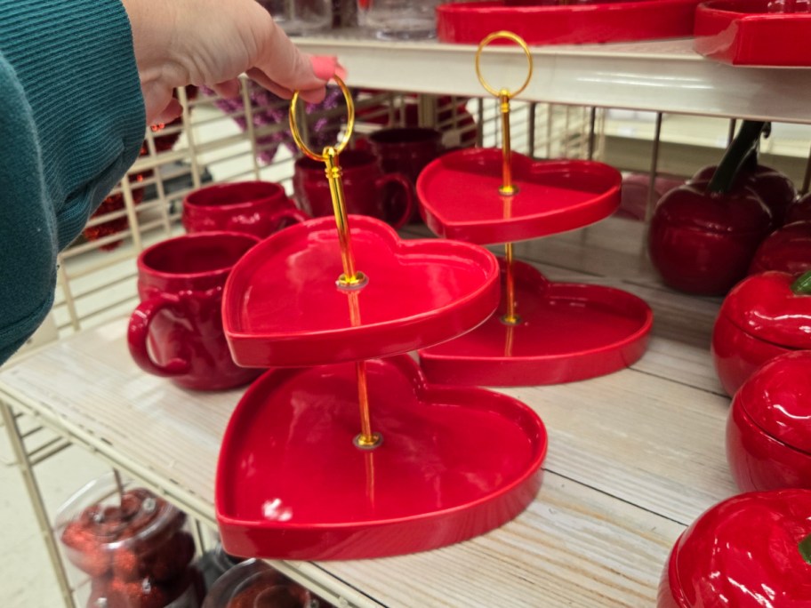 A hand holds a two-tiered heart-shaped serving tray with a gold handle. The vibrant red trays are set against shelves filled with matching red dishes.