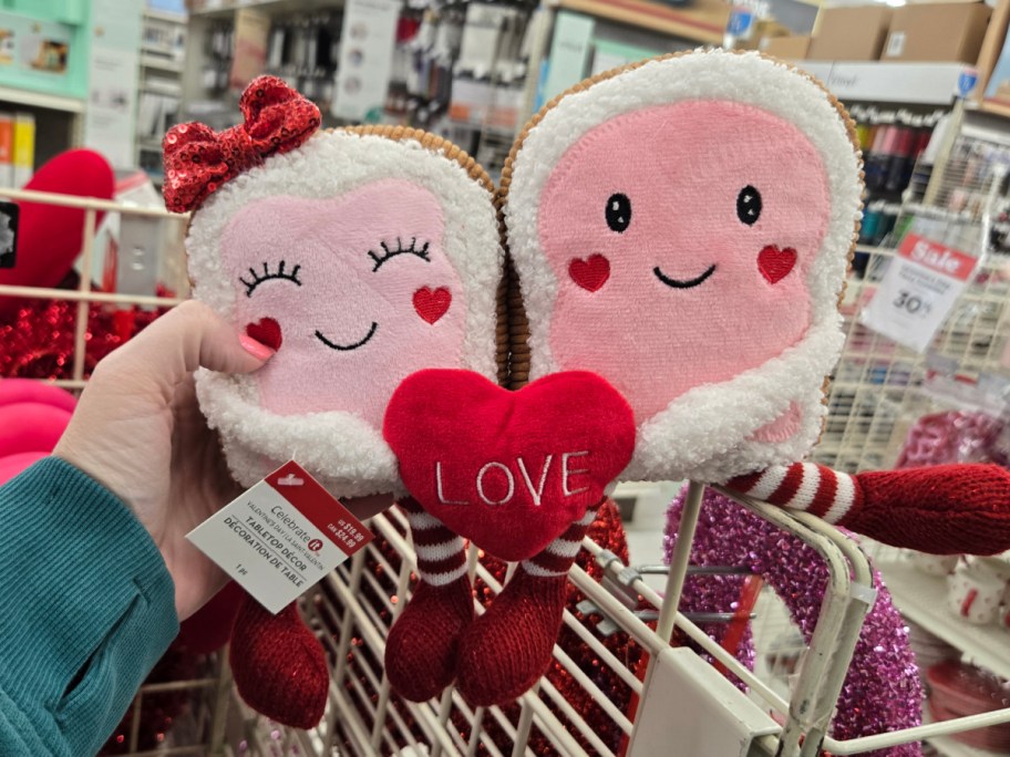 Two cute plush toast figures with smiling faces and red heart cheeks are held in hand. They hold a red heart labeled "LOVE" in a store setting.