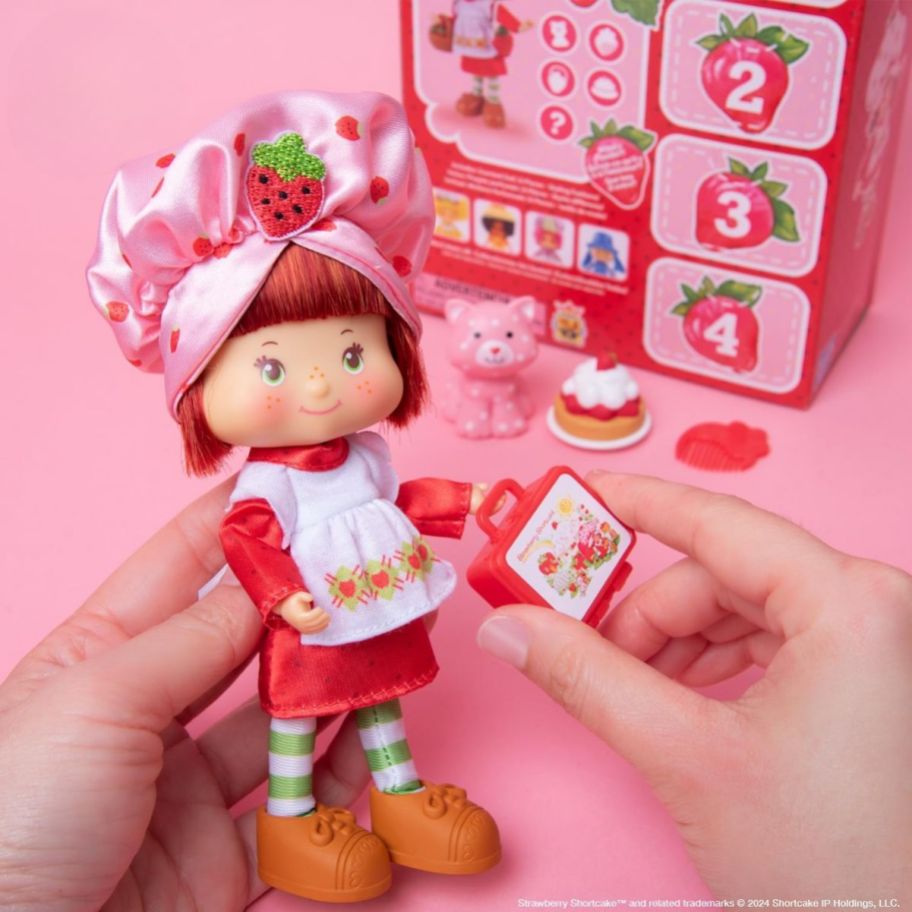 a pair of hands holding a strawberry shortcake doll, with accessories in the background