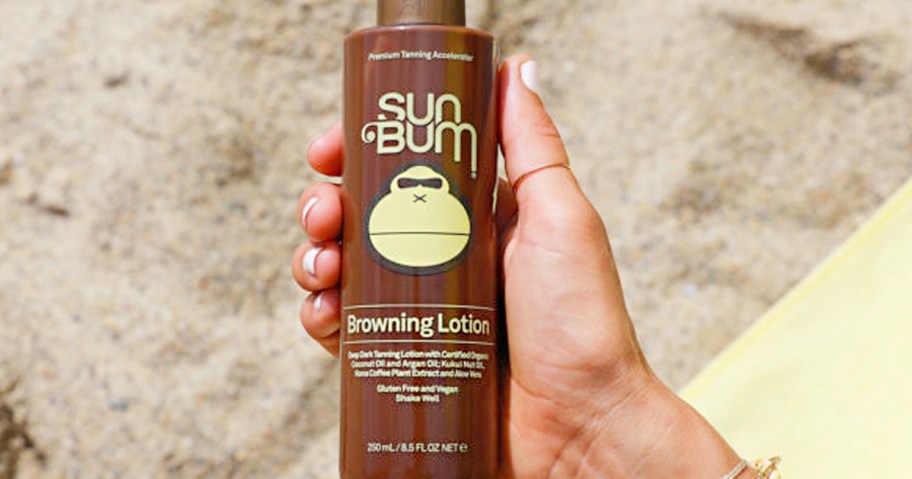 hand holding sun bum browning lotion bottle over beach sand