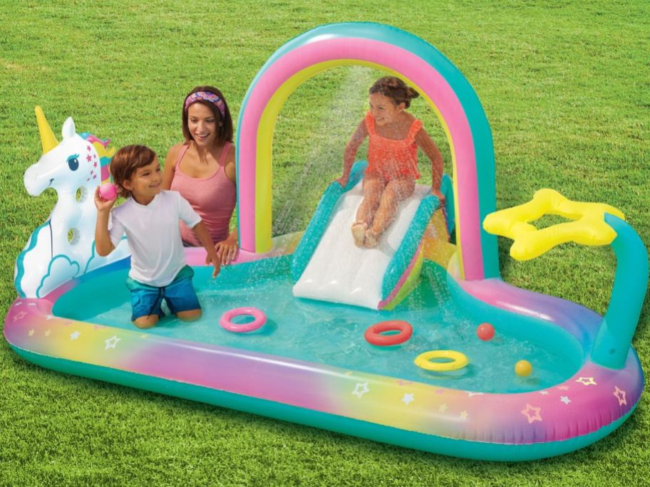 two kids playing in an inflatable rainbow unicorn theme pool with a mom sitting nearby