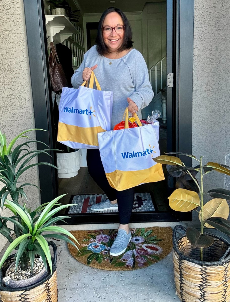 smiling woman standing on her doorstep with the door open, holding two walmart plus grocery delivery bags
