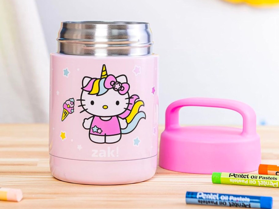 pink hello kitty food jar with pink lid laying on table with crayons