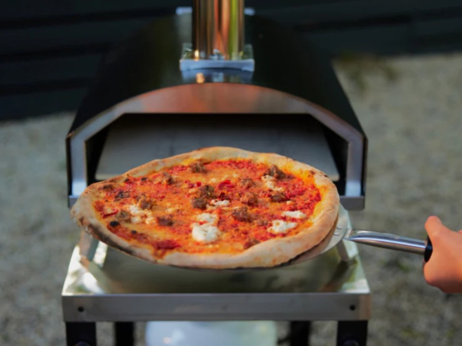 person pulling a pizza out of a black and steel outdoor wood pizza oven