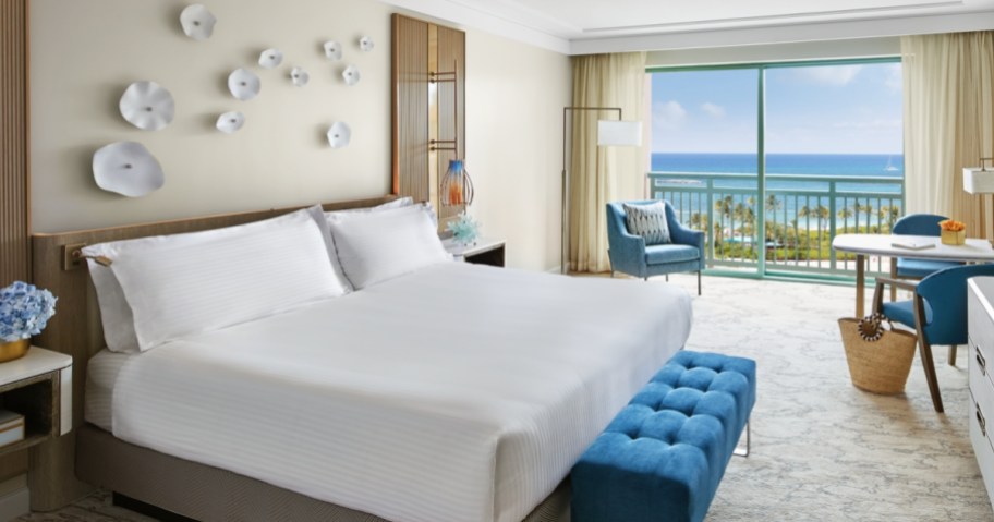 Atlantis Bahamas resort hotel room with large king bed with white bedding, blue bench and chair, ocean view from the balcony in the background