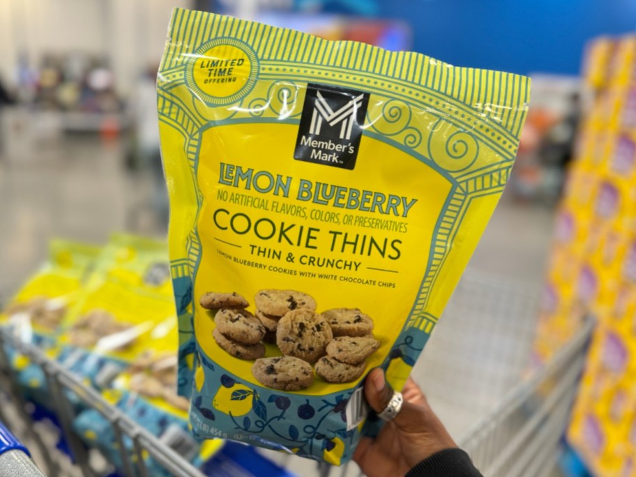 hand holding a bag of Member's Mark Lemon Blueberry Cookie Thins with cart with more bags in it