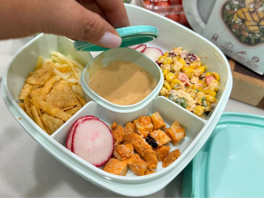 A Bentgo salad container with all of the compartments filled with salad ingredients and a hand lifting the lid off the dressing compartment in the middle