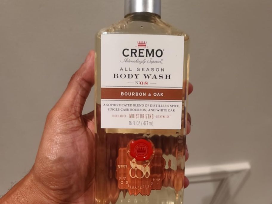 A hand holding a bottle of Cremo Body Wash Bourbon & Oak