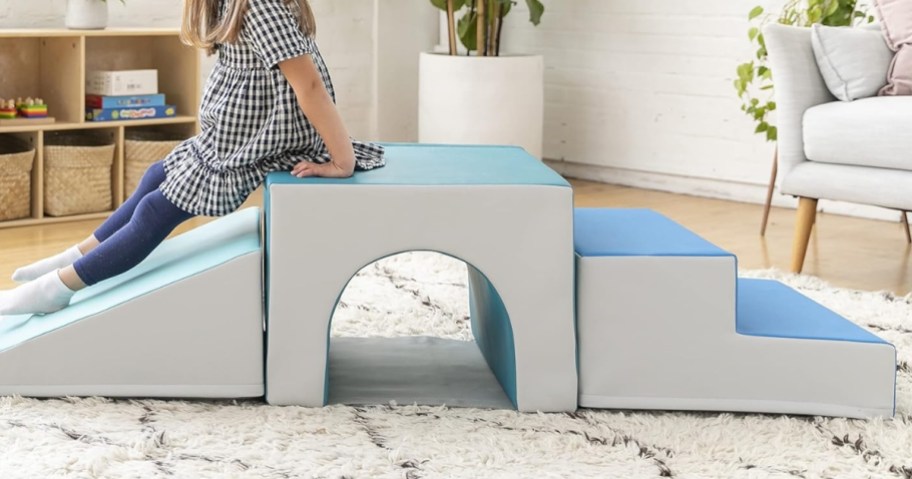 little girl sliding on a kid's tunnel and step playset that's set up in a living room