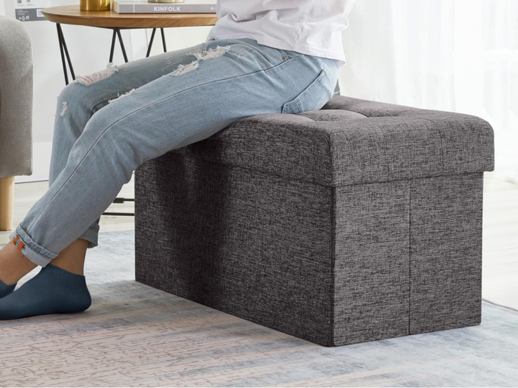 Person in blue jeans sitting on gray ottoman next to small table