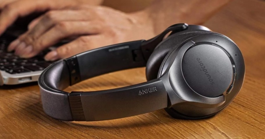 black pair of Anker headphones laying on desk, person's hand on a laptop behind them