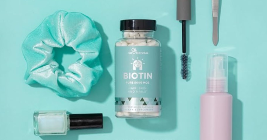 bottle of Biotin supplements surrounded by a hair scrunchie, nail polish, mascara want and spray bottle on a green background