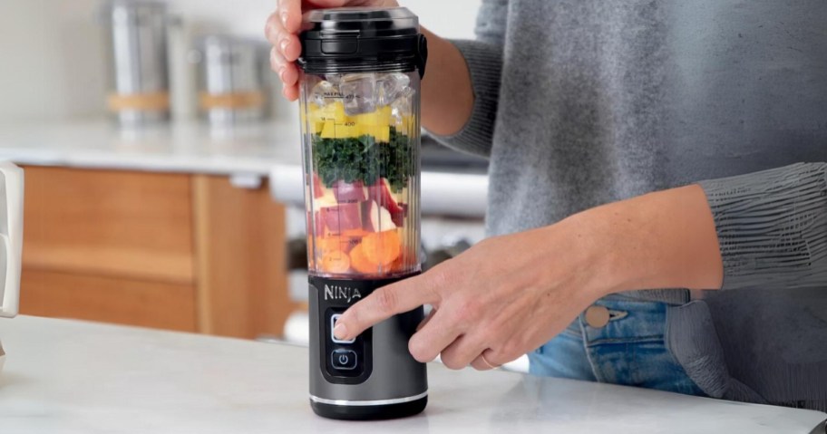 Ninja personal blender with produce inside, person pushing the button to blend the food and holding the top with their other hand