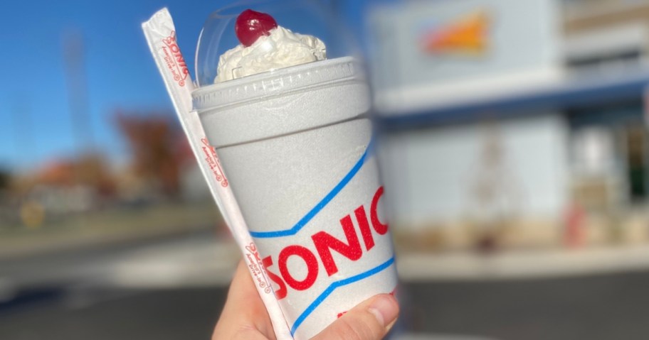 hand holding a Sonic ice cream shake in a sonic cup with a straw, Sonic Drive in in the background