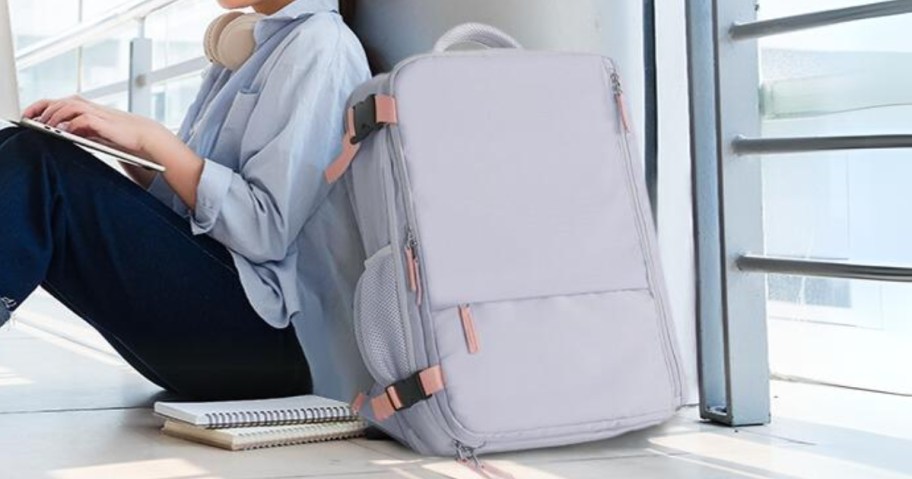 girl sitting on the floor at an airport, large light purple and pink travel backpack leaned against the wall beside her